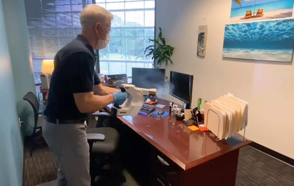 Professional cleaner wearing gloves and a face mask disinfecting an office desk and computer equipment with an electrostatic sprayer.